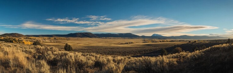 Obraz premium A vast valley stretches under a clear blue sky filled with wispy clouds. Sunlight bathes the grassy terrain and distant mountains as autumn foliage adds color