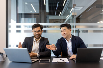 Business meeting, two men sitting inside office with laptops, smiling cheerfully discussing work...