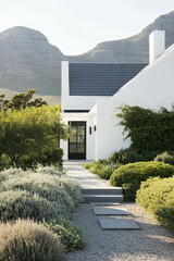 Modern white house with black accents nestled in a serene garden landscape under a clear blue sky surrounded by majestic mountains