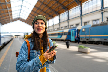 A female tourist in a train station, holding her phone and carrying a backpack, ready to embark on...