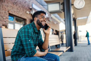 Young smiling black man sitting at train station and having phone call while waiting for train.