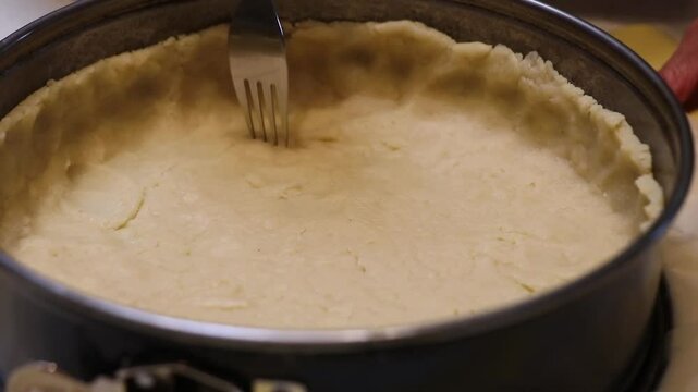 A round metal springform pan holds an uncooked dough crust with fork piercings. A hand steadies the pan on a light colored countertop during baking prep.