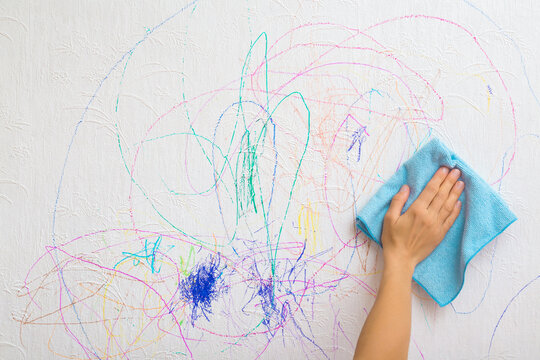 Woman hand holding blue microfiber rag and wiping scratches lines of colorful markers from white wallpaper surface. Mother cleaning wall from draw of naughty little kid. Closeup. Front view.