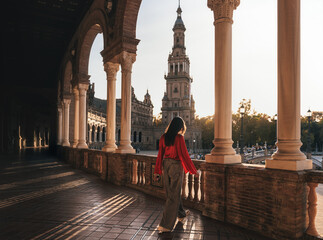 Walks along the colonnaded walkway of Plaza de España, Seville, Spain. The iconic tower and ornate columns are illuminated by golden sunset light, showcasing historic architecture.