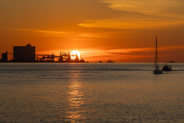 Fototapeta premium Vibrant sunset over Tagus River in Lisbon, Portugal, featuring dock silhouettes and sailboats in a calm and orange-toned seascape