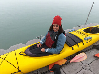 Obraz premium Young brunette woman smiling at the camera while seated in a yellow kayak, ready to paddle in cold waters. Dressed warmly for outdoor adventure on a calm, overcast day