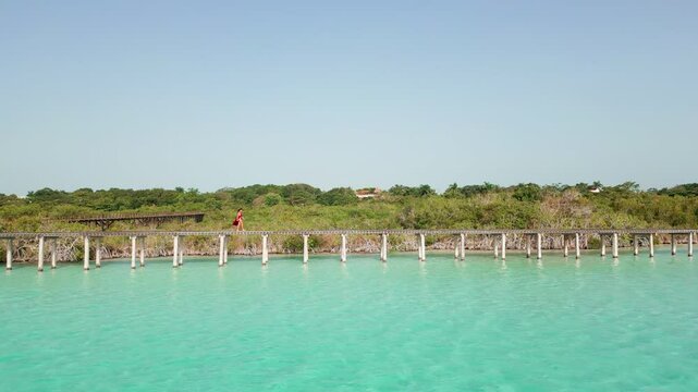 Toma a&eacute;rea cenital de dron de joven y atractiva mujer en vestido rojo, caminando en un muelle de madera, en el agua color turquesa y los manglares del Parque Ecol&oacute;gico de Bacalar en M&eacute;xico.