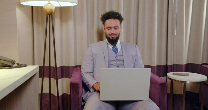 In a luxurious hotel room, a sharply dressed man unwinds while focusing on his laptop. The contemporary workspace inspires admiration and aspiration in those wishing to emulate this lifestyle