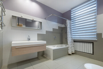 The bathroom is decorated in light gray tones, with an elegant interior design. A bathtub with a shower behind a curtain, blinds on the window.