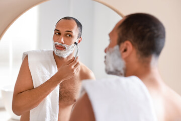 Handsome man shaving with razor near mirror in bathroom