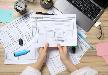 Website designer holding wireframe at wooden table with stationery and laptop, top view