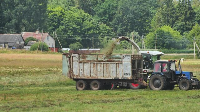 Rural tractor unloading golden hay bales into trailer, sunlight illuminating agricultural landscape near traditional village