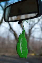 car's rearview mirror with a green leaf-shaped air freshener hanging from it bare trees...