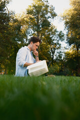A man sitting on the grass in a park, studying with a book in hand, surrounded by greenery,...
