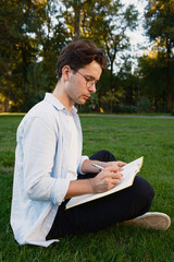 A young man wearing glasses and casual clothing is seated on a grassy area in a park, writing in a...
