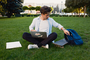 Fototapeta premium A student sits cross-legged on grass in a park, using a laptop, with a skateboard and a backpack beside him, suggesting relaxation and learning.