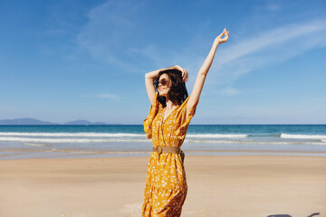 Joyful woman in a vibrant yellow dress with arms raised on the sandy beach at sunset, feeling free and happy