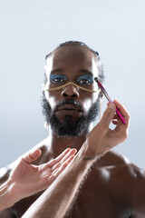 Crop hands of makeup artist applying eye makeup on African American gay model over gray background. Portrait of serious homosexual man with chain on nose is in studio. 