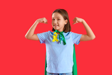 Little girl dressed as superhero with gold ribbon showing muscles on red background. Childhood cancer awareness concept