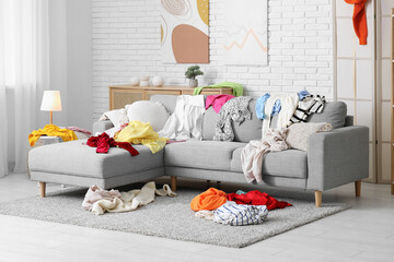 Interior of messy living room with grey sofa and piles of different scattered clothes