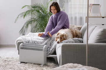 Young woman with blanket and Australian Shepherd dog sitting on sofa near heater at home