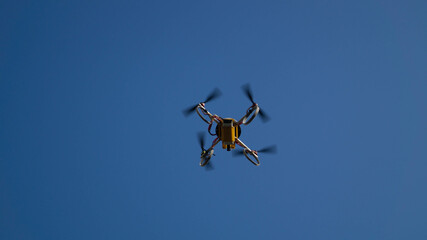 A low-angle, full shot captures a small, yellow drone in flight against a clear, vibrant blue sky.
