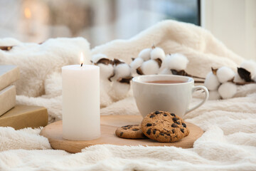 Burning candle with tea cup and cookies on plaid near window, closeup