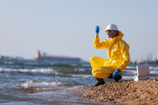 A person wearing a yellow protective hazmat suit and white helmet kneeling on a sandy shore, holding a water sample vial near the ocean. A large cargo ship is visible in the background