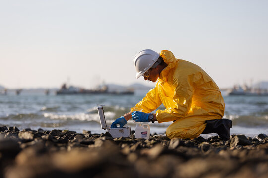 A person wearing a yellow hazmat suit and white helmet kneeling on a rocky shore,performing water quality testing using a sample vial and a testing kit. The sea and boats are visible in the background