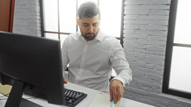 Young man working in a modern office, taking notes at his desk with a computer in front, showcasing a focused and professional atmosphere.