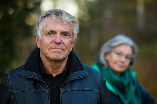A portrait of an older Swedish man looking into the camera and his wife out of focus in the background