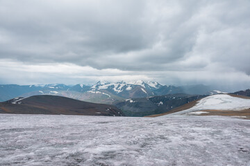 Dramatic panoramic view from big glacier to wide alpine valley and large snow-capped mountain range in rainy low clouds. Awesome vast landscape with high snowy mountains in rain under gray cloudy sky.
