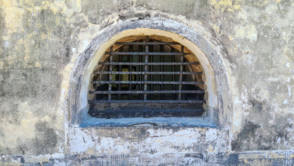 Fototapeta premium A barred arched window in a weathered stone wall evokes a sense of confinement, history, and the passage of time