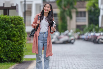 Happy asian female college student walking through campus, holding books and carrying a backpack, smiling brightly while looking away, embracing the joy of education and youth