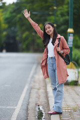 Happy smiling young asian woman student waving hand hailing taxi cab while standing on sidewalk on city street commuting to work or university