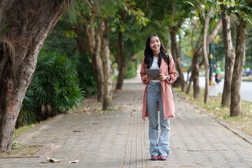 Young asian woman walking in a tree-lined park, holding a digital tablet and wearing casual clothing, enjoying the pleasant surroundings during her commute