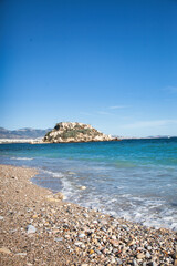 Obraz premium Picturesque pebble beach with turquoise Mediterranean waters and rocky island in Athens, Greece. Crystal clear sea waves washing onto stone-covered shore under bright blue summer sky.