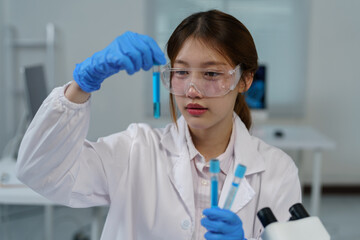 Young female scientist wearing lab coat and protective glasses, carefully examining test tubes containing blue liquid, conducting experiments in modern laboratory