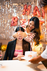 Joyful asian lady surprises her friend playfully covering eyes before giving birthday cake, celebrating special moment together in house part. Holiday Celebration, Birthday.