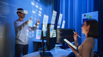 developer uses a VR headset to interact with projected interface designs, while colleagues discuss...