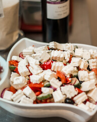 A vibrant salad with cucumbers, tomatoes, red onions, olives, and feta cheese in a white bowl. A wine bottle and kitchen items are visible in the background.