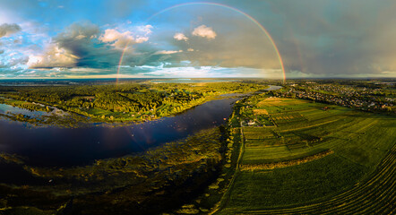 Vibrant aerial view of a rural area featuring a wide river, fields, forests, a rainbow with a faint secondary arc, and scattered houses under a cloudy sky.