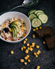 A white bowl holds a mixed salad with cucumbers, corn, red onion, and a black olive on a dark surface, surrounded by bread and scattered ingredients.
