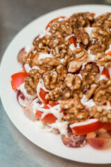 A white plate on a metallic surface holds halved walnuts, sliced red tomatoes, and grapes, topped with a drizzle of creamy white dressing.