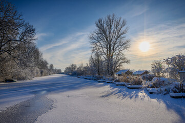 Winter scene with frozen river covered in snow, surrounded by frost-covered trees, wooden berths, boat and huts. Eider river near Kluvensiek locks in winter, Rendsburg, Schleswig-Holstein, Germany.