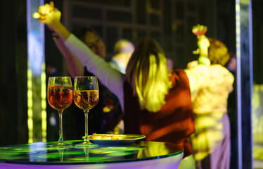 Obraz premium Colorful party scene with wine glasses in foreground, friends dancing and celebrating in vibrant lighting.
