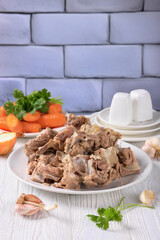 pork and beef bones with leftover meat after cooking broth on a white plate with carrot, garlic, onion and spices at the background of brick wall, vertical view from above