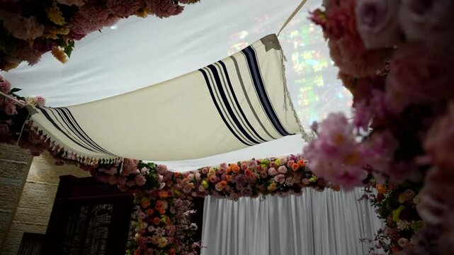 Beautiful indoor chuppah adorned with vibrant pink and orange roses, white fabric canopy, and floral details. Set against a stone wall, perfect for Jewish wedding themes and event inspiration