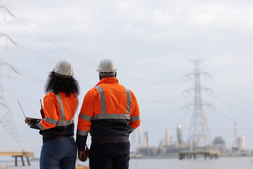 Two engineers in orange safety jackets and helmets stand facing a power plant in the distance,...