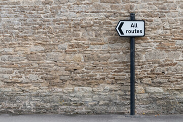All Routes Road Sign pointing left in front of an old stone wall